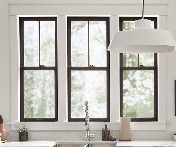 Three black-framed double-hung windows above a white kitchen counter looking out to trees