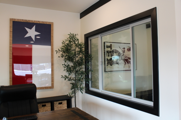 Black-framed horizontal slider window installed in an interior wall beside a framed Texas flag