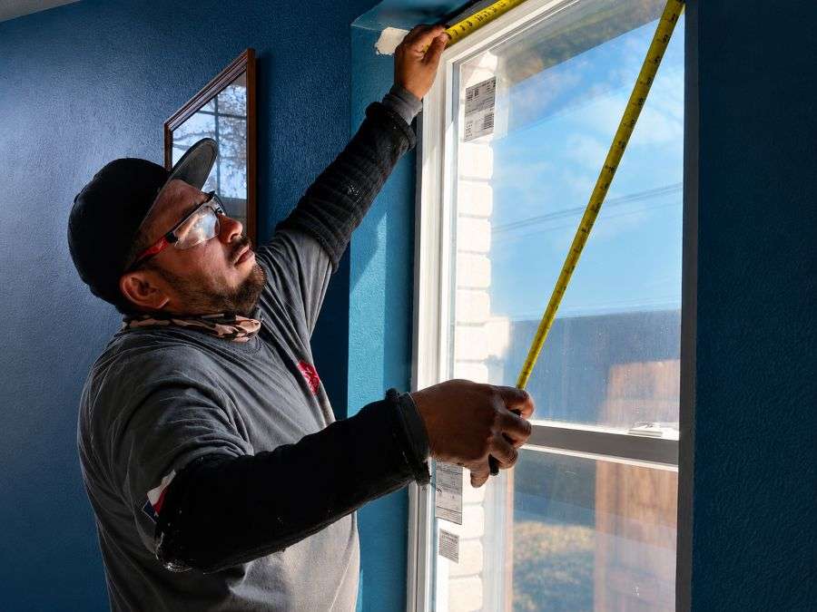 Hardy Windows of Texas professional measures a white replacement window with a yellow tape measure indoors.